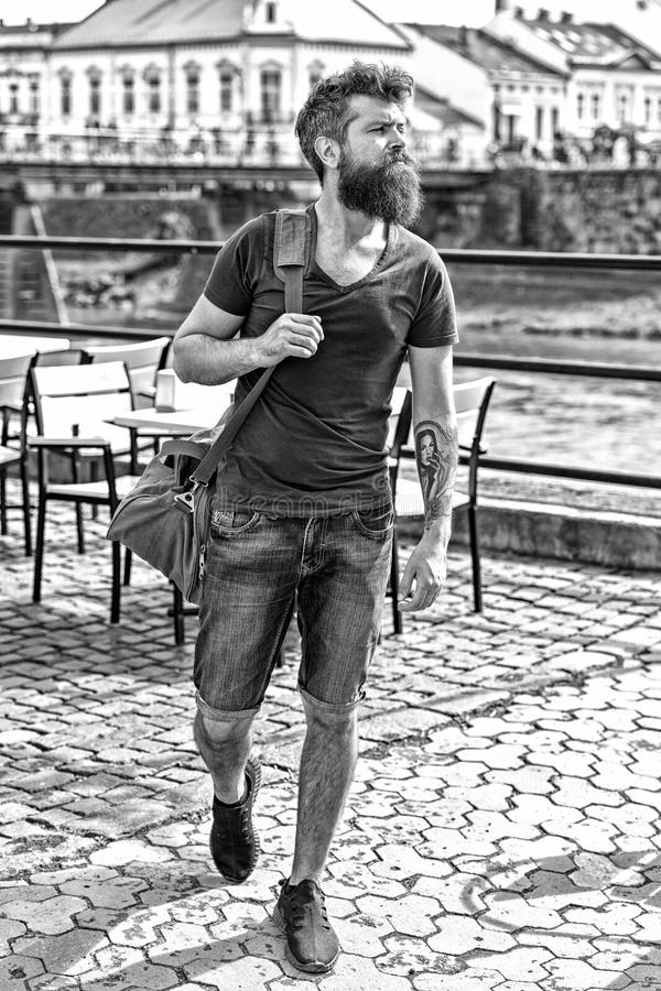 Homem muscular com barba à moda e bigode que leva o saco desportivo grande ao andar na cidade velha Homem novo na sarja de Nimes imagem de stock royalty free Homem muscular com barba à moda e bigode que leva o saco desportivo grande ao andar na cidade velha Homem novo na sarja de Nimes imagem de stock royalty free