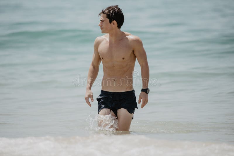 Homem muscular considerável novo que anda fora da água em uma praia tropical que veste um maiô