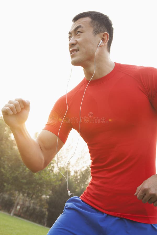Young muscular man with a red shirt running and listening to music on ear buds outdoors in the park in Beijing, China, with lens flare. Young muscular man with a red shirt running and listening to music on ear buds outdoors in the park in Beijing, China, with lens flare