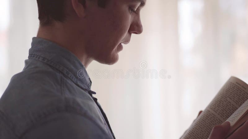 Homem novo adorável do retrato que lê livro interessante na sala na frente do grande fim da janela acima Lazer em casa