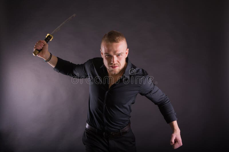 Homem novo agressivo que guarda a espada do katana que olha a câmera Contra o fundo da parede foto de stock Homem novo agressivo que guarda a espada do katana que olha a câmera Contra o fundo da parede foto de stock