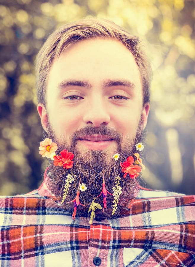Homem novo com uma barba longa e flores em sua barba em um fim borrado natural vermelho do fundo da camisa de manta acima imagens de stock royalty free Homem novo com uma barba longa e flores em sua barba em um fim borrado natural vermelho do fundo da camisa de manta acima imagens de stock royalty free