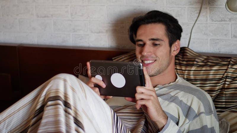 Homem novo considerável na cama que datilografa no PC da tabuleta