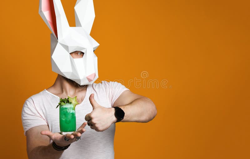 Homem novo na máscara de papel do coelho e no t-shirt branco que bebe a bebida verde do cocktail foto de stock Homem novo na máscara de papel do coelho e no t-shirt branco que bebe a bebida verde do cocktail foto de stock