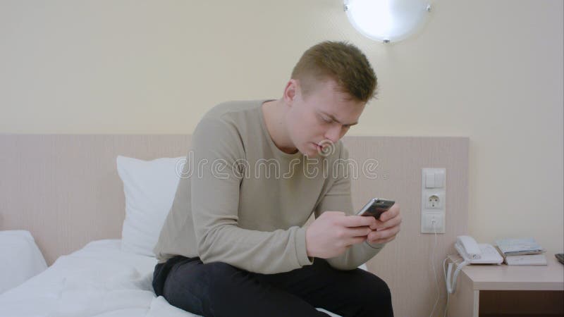 Homem novo preocupado que datilografa uma mensagem dos sms em seu telefone celular que senta-se na cama em casa foto de stock Homem novo preocupado que datilografa uma mensagem dos sms em seu telefone celular que senta-se na cama em casa foto de stock