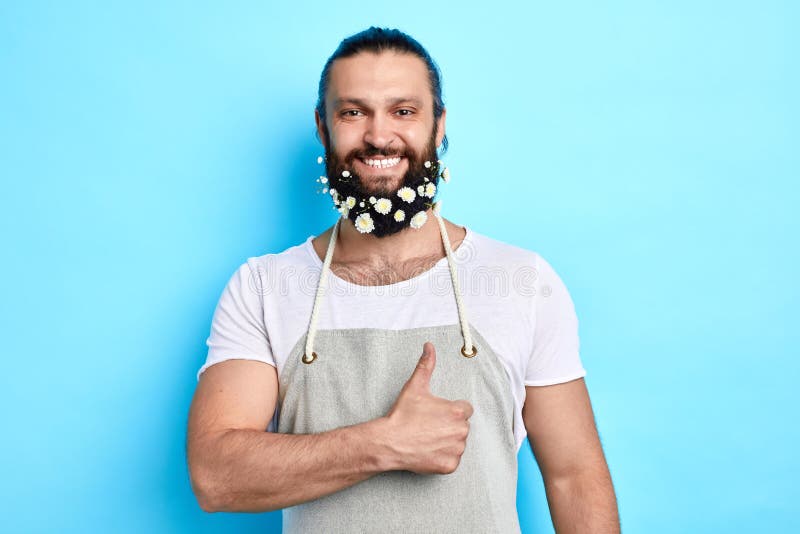 Homem positivo alegre com a barba decorada com as flores que ahowing o polegar acima foto de stock Homem positivo alegre com a barba decorada com as flores que ahowing o polegar acima foto de stock