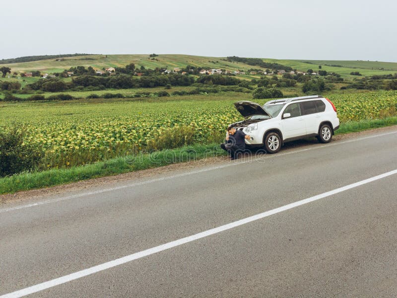 Homem que senta-se na estrada perto de carro quebrado carro de tentativa da parada para a ajuda imagem de stock Homem que senta-se na estrada perto de carro quebrado carro de tentativa da parada para a ajuda imagem de stock