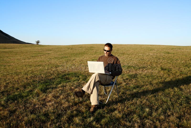 Homem que senta-se no campo com portátil imagem de stock Homem que senta-se no campo com portátil imagem de stock
