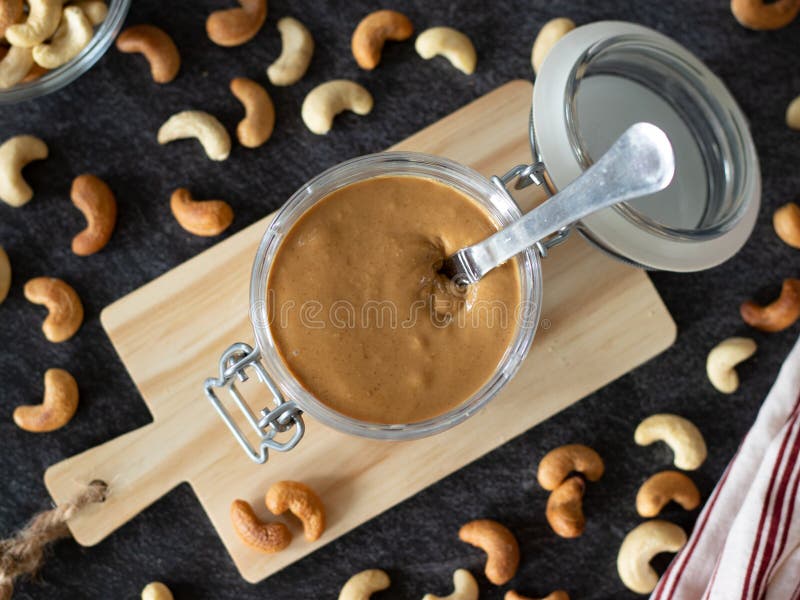 Homemade cashew butter in a jar with roasted and raw cashew nuts on dark background