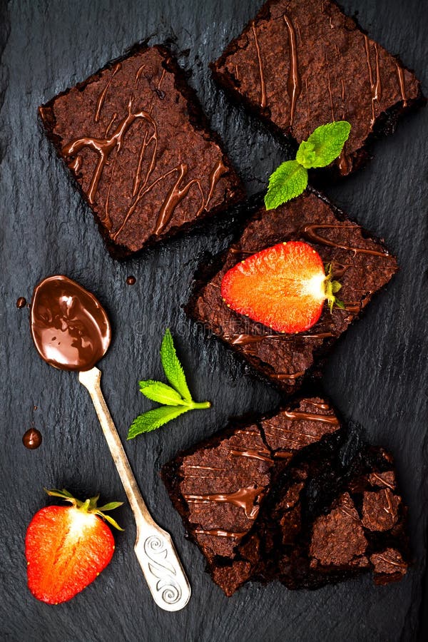 Homemade dark chocolate brownies decorated with strawberries and mint leaves over black slate background, top view.