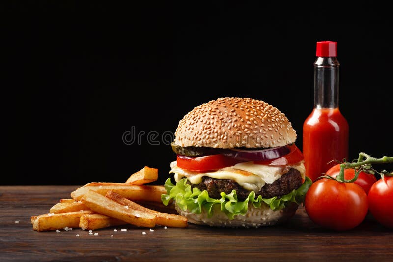 Homemade hamburger close-up with beef, tomato, lettuce, cheese, french fries sauce bottle on wooden table. Fastfood on dark