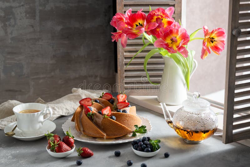 Homemade sponge cake with fresh berries on the table with tulips and tea pot. Side view. Bakery, pastry, recipe, poster, greeting