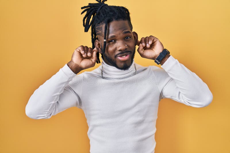Homme africain avec des poignées de rechapage portant un pull à col roulé sur fond jaune sourire tirant oreilles avec les doigts geste drôle. problème d'audition