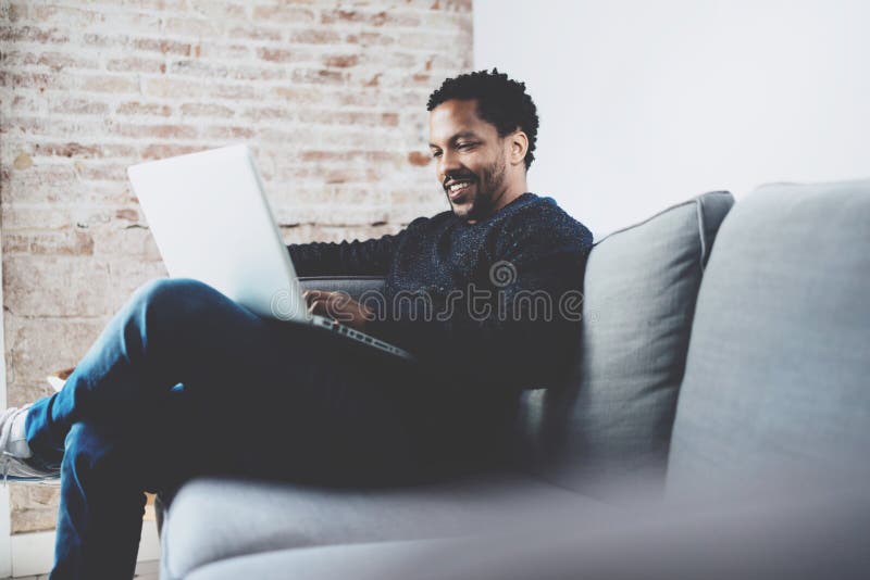 Homme africain barbu gai travaillant sur l'ordinateur portable tout en reposant le sofa à son endroit moderne de bureau Concept d image stock Homme africain barbu gai travaillant sur l'ordinateur portable tout en reposant le sofa à son endroit moderne de bureau Concept d image stock
