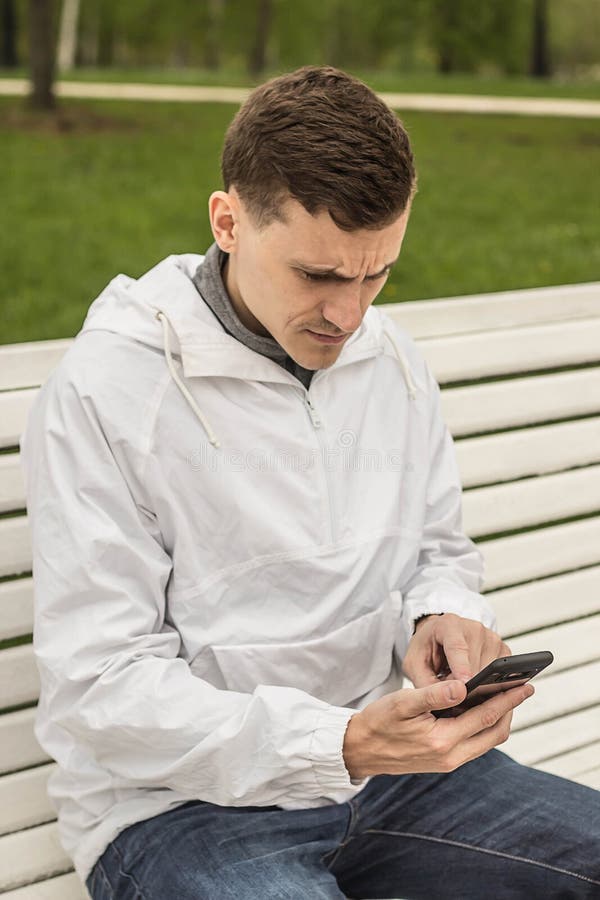 Homme avec le chapeau se reposant sur un banc en bois et vérifiant le message par l'écran de smartphone avec une expression série image stock Homme avec le chapeau se reposant sur un banc en bois et vérifiant le message par l'écran de smartphone avec une expression série image stock