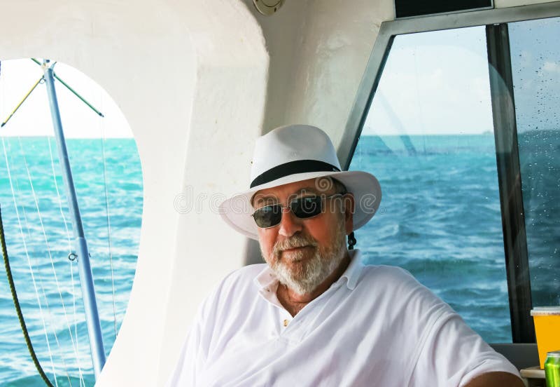 Homme barbu d'une chevelure gris avec le chapeau détendant sur le bateau profond de pêche maritime avec l'océan à l'arrière-plan images stock Homme barbu d'une chevelure gris avec le chapeau détendant sur le bateau profond de pêche maritime avec l'océan à l'arrière-plan images stock