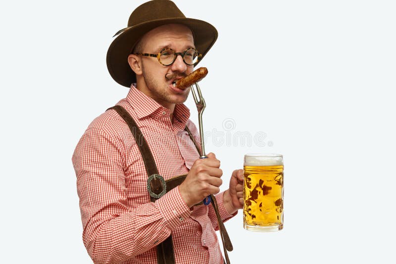 Attractive bavarian man wearing traditional fest outfit holding huge beer mug and bites hot fried sausage. Concept of Oktoberfest, traditions, drinks and food. Copy space for ad. Attractive bavarian man wearing traditional fest outfit holding huge beer mug and bites hot fried sausage. Concept of Oktoberfest, traditions, drinks and food. Copy space for ad