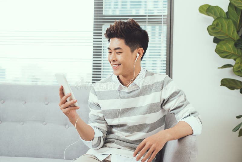 Homme bel asiatique de sourire s'asseyant sur le sofa confortable écoutant le mus photo stock Homme bel asiatique de sourire s'asseyant sur le sofa confortable écoutant le mus photo stock