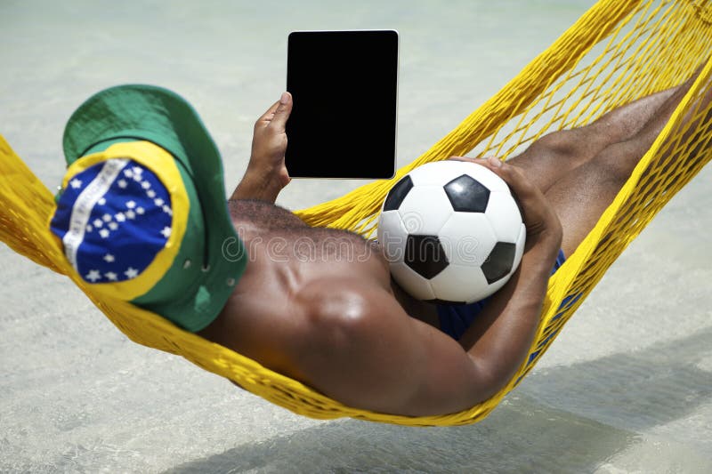 Homme brésilien détendant avec l'hamac de Tablette et de plage du football photographie stock Homme brésilien détendant avec l'hamac de Tablette et de plage du football photographie stock