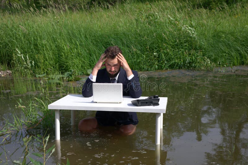 Tired, depressed, boring, bearded businessman disappointed with a failed business deal, looking at a laptop. having headache. At the table, standing in the swamp, in the water. Tired, depressed, boring, bearded businessman disappointed with a failed business deal, looking at a laptop. having headache. At the table, standing in the swamp, in the water