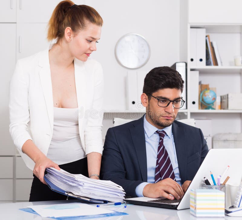 Businesswoman bringing to unhappy male coworker big pile of files. Businesswoman bringing to unhappy male coworker big pile of files