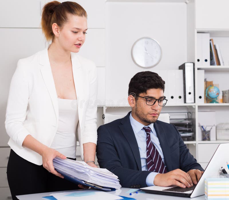Businesswoman bringing to unhappy male coworker big pile of files. Businesswoman bringing to unhappy male coworker big pile of files