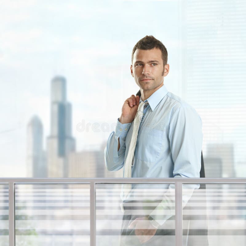 Casual businessman standing in front of windows in office, smiling. Skyscrapers in background. Casual businessman standing in front of windows in office, smiling. Skyscrapers in background.