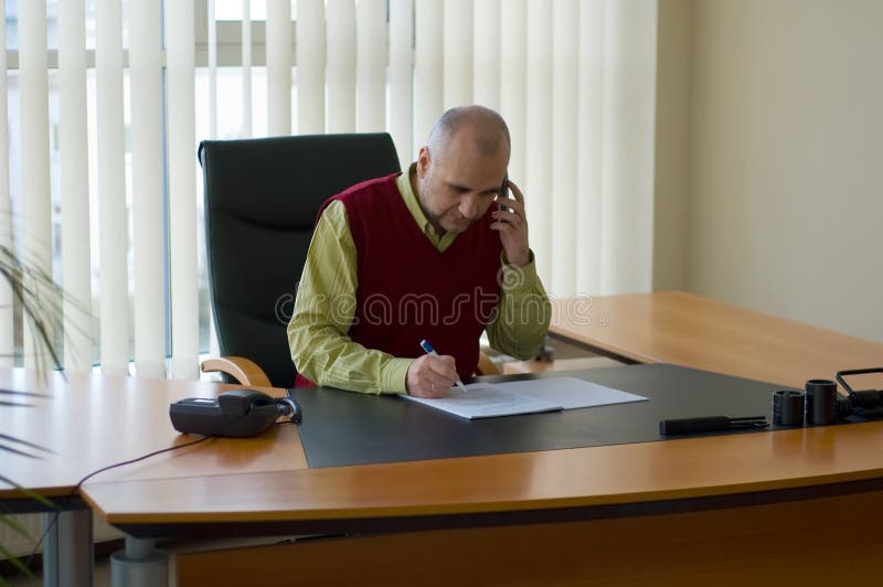 Casual businessman studying agreement at his desk by window and speaking on the cellphone. Casual businessman studying agreement at his desk by window and speaking on the cellphone