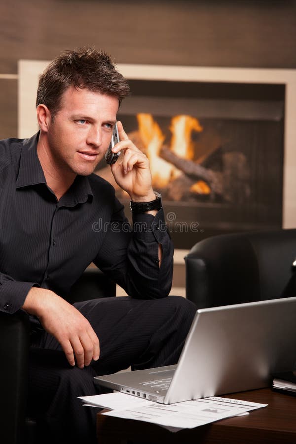 Casual businessman sitting in armchair at coffee table in front of fireplace, talking on phone. . Casual businessman sitting in armchair at coffee table in front of fireplace, talking on phone. .