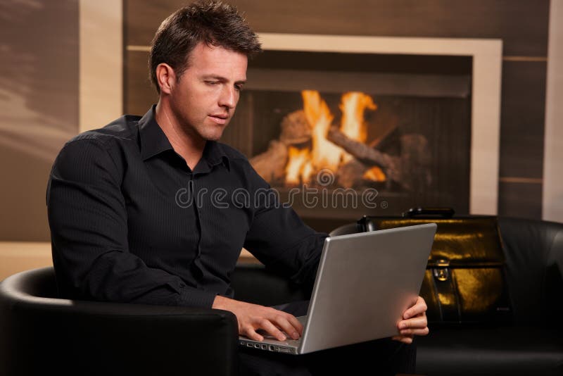 Casual businessman using laptop computer at home, sitting in armchair in front of fireplace. Casual businessman using laptop computer at home, sitting in armchair in front of fireplace.