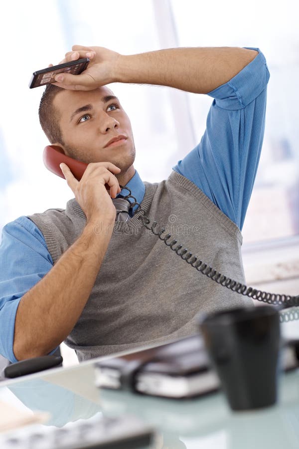Troubled businessman at desk concentrating on landline phone call, holding mobile phone, looking up. Troubled businessman at desk concentrating on landline phone call, holding mobile phone, looking up.