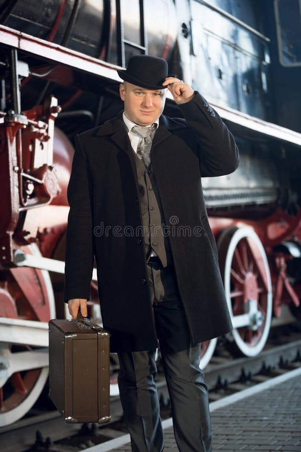Homme dans le rétro costume, le chapeau de lanceur et la valise posant au vieux locomot image stock Homme dans le rétro costume, le chapeau de lanceur et la valise posant au vieux locomot image stock
