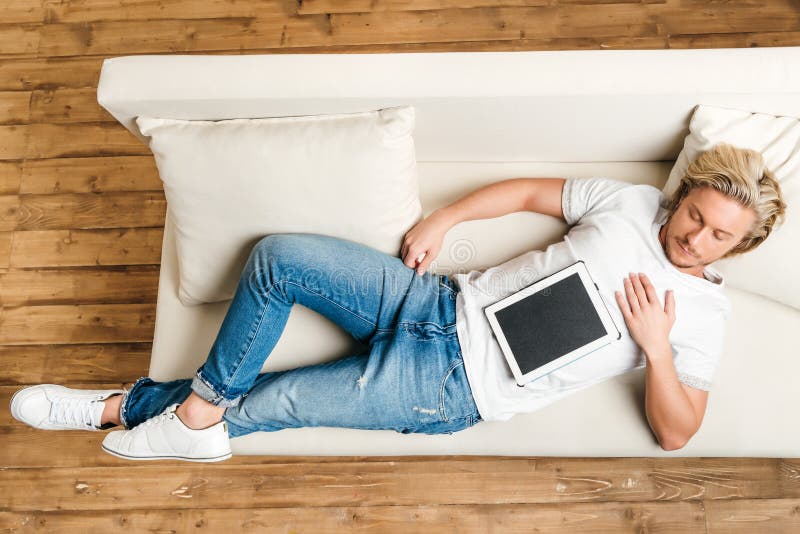 Homme dans le T-shirt et des jeans blancs dormant sur le sofa avec le comprimé numérique photographie stock libre de droits