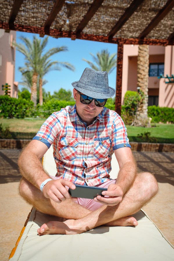 Homme dans les lunettes de soleil et un chapeau reposant sur un canapé et jeux dans le smartphone photo stock Homme dans les lunettes de soleil et un chapeau reposant sur un canapé et jeux dans le smartphone photo stock