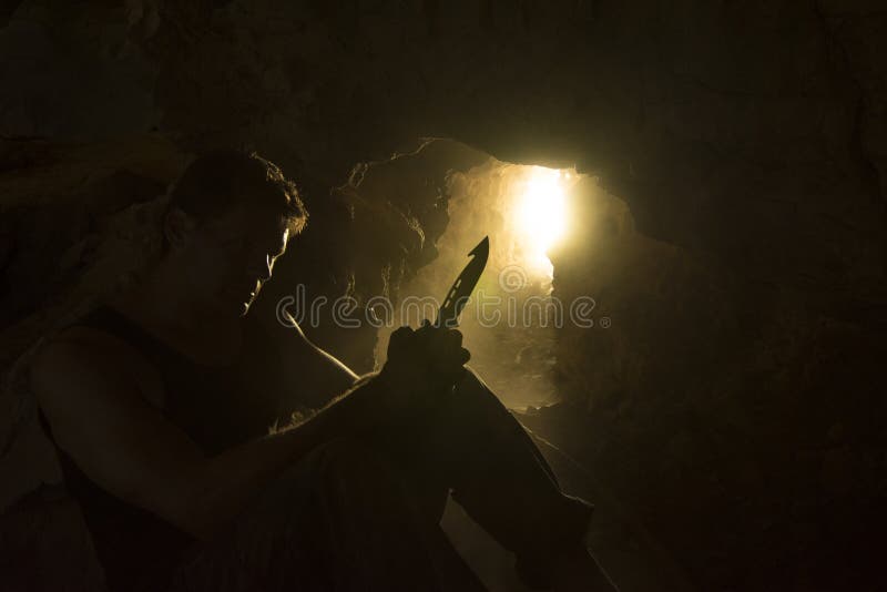 Silhouette with rim light of adventure man sitting in dusty cave looking at and holding survival knife. Silhouette with rim light of adventure man sitting in dusty cave looking at and holding survival knife