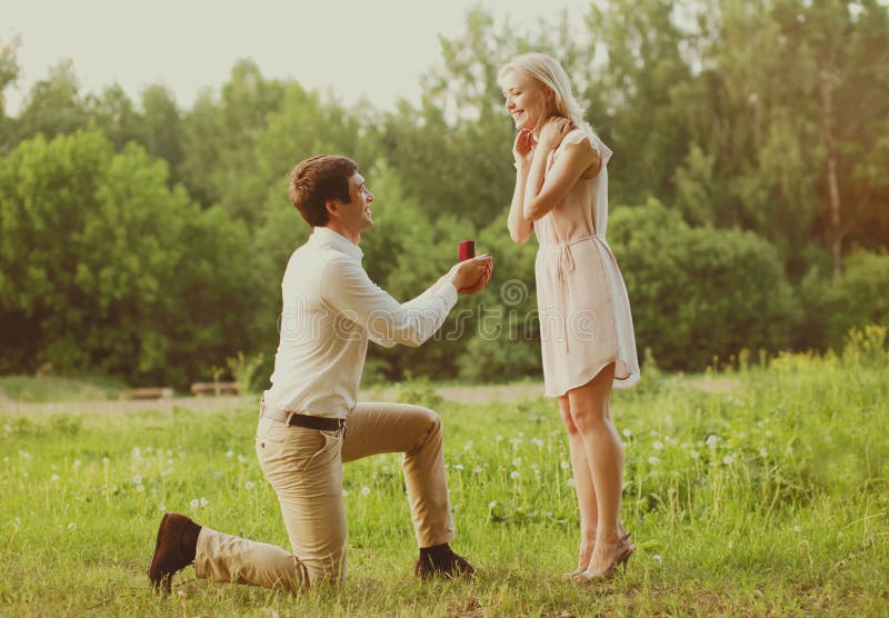 Homme heureux de couples se mettre à genoux et proposant l'anneau à sa femme dehors sur le concept de mariage d'herbe images stock