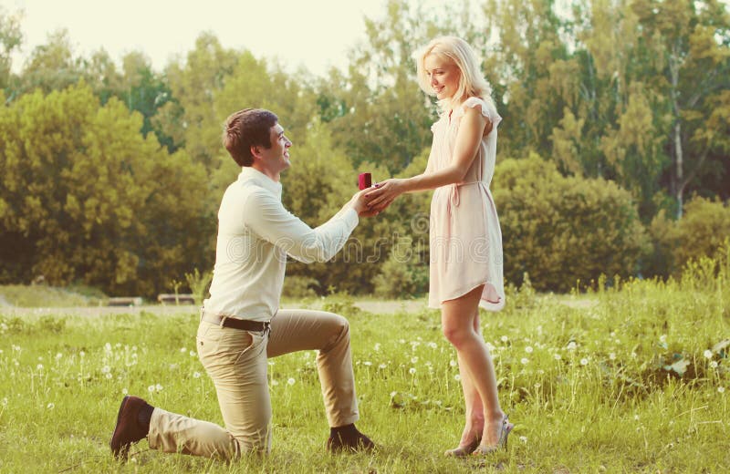 Homme heureux de couples se mettre à genoux et proposant l'anneau à sa femme dehors sur le concept de mariage d'herbe photo stock