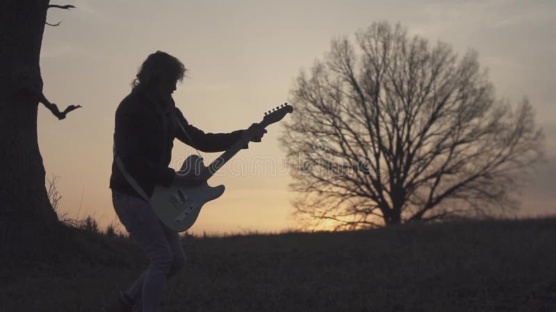 Homme jouant la guitare électrique et chantant dans un domaine près de l'arbre au coucher du soleil Silhouette