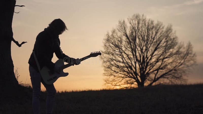 Homme jouant la guitare électrique et chantant dans un domaine près de l'arbre au coucher du soleil Silhouette