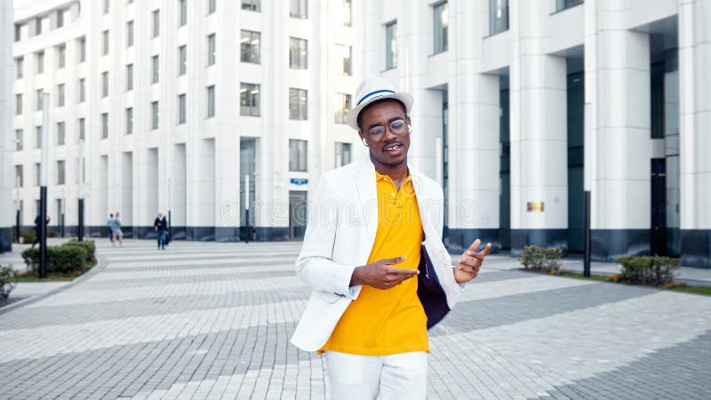 Joyful black man in glasses and white wireless headphones listens to music and dances walking along grey pavement in megalopolis. Joyful black man in glasses and white wireless headphones listens to music and dances walking along grey pavement in megalopolis