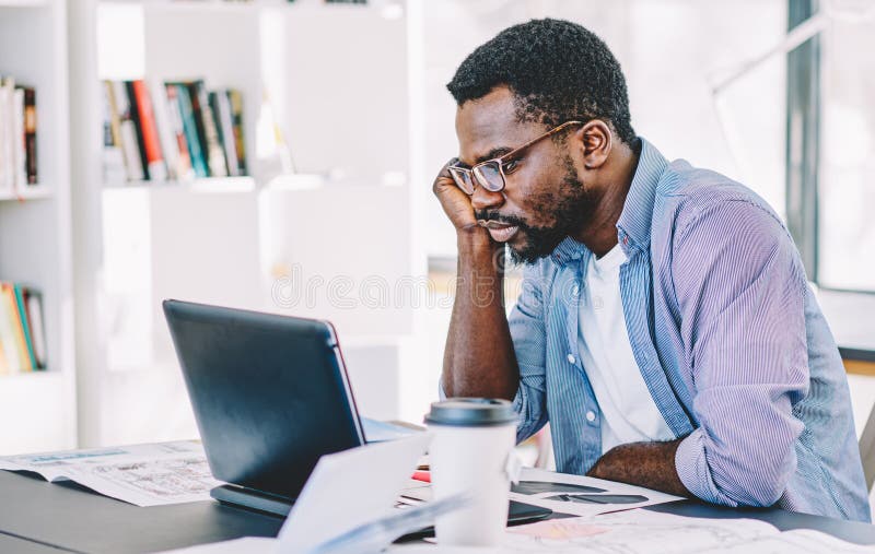 African American man in glasses wearing shirt browsing on laptop sitting at table loaded with glasses notebooks markers in office space. African American man in glasses wearing shirt browsing on laptop sitting at table loaded with glasses notebooks markers in office space