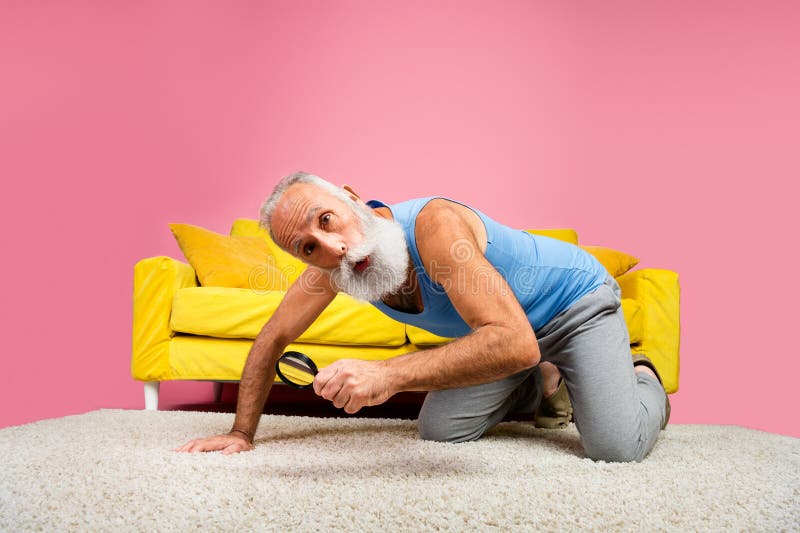 Homme senior avec une loupe en verre regardant sous les meubles dans un salon lumineux avec mur rose et décoration vibrante photos stock