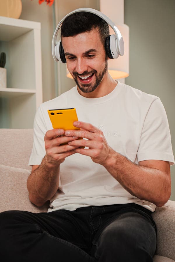 Homme souriant vertical portant un casque à l'aide d'un smartphone jaune à la maison en écoutant de la musique ou des textos assis photo libre de droits