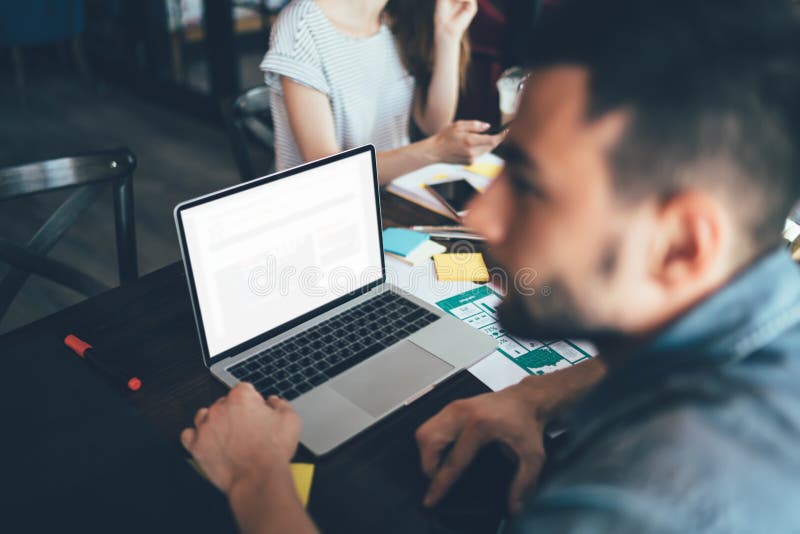 Young bearded male in casual jeans apparel watching laptop sitting at wooden table loaded with documents stickers smartphone in office. Young bearded male in casual jeans apparel watching laptop sitting at wooden table loaded with documents stickers smartphone in office