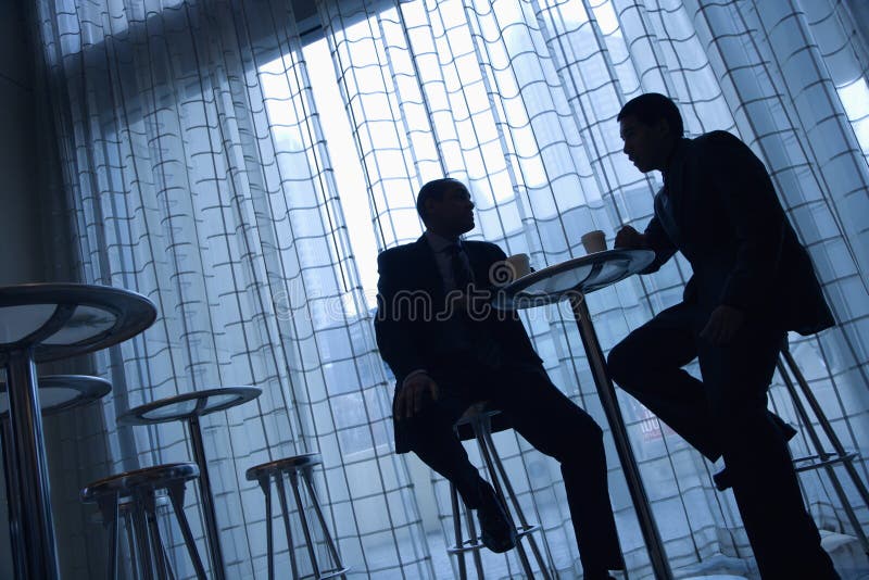 Tilt view silhouette of African-American and Asian businessmen sitting at a table having coffee in front of a curtained window. Tilt view silhouette of African-American and Asian businessmen sitting at a table having coffee in front of a curtained window.