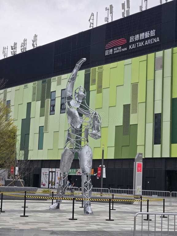 A large metallic sculpture of two athletes in a sports pose is placed in front of the Kai Tak Sports Park building, which features a modern green and black facade with signage indicating it is the Kai Tak Arena.