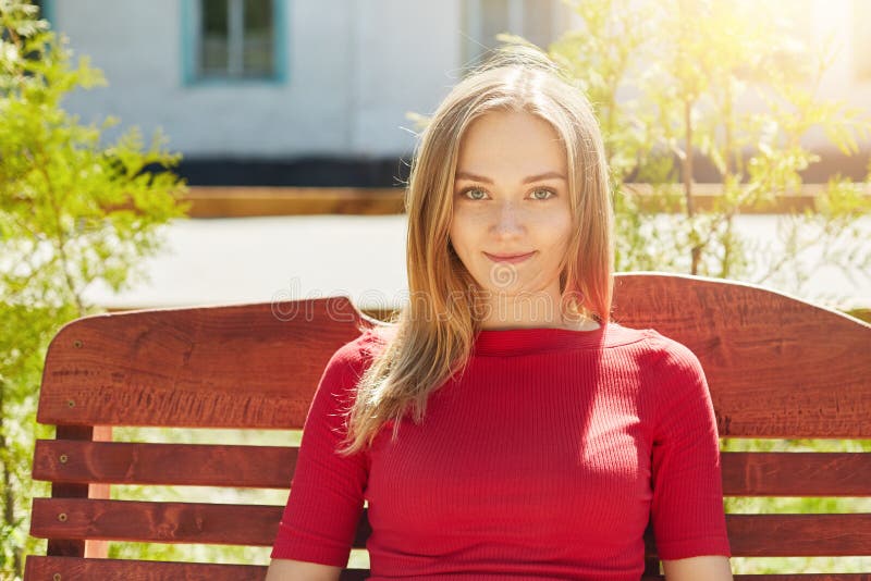 Horizontal portrait of fair-haired pretty young girl with freckles and blue eyes wearing red sweater sitting at bench in park admi