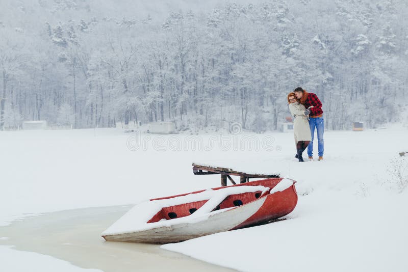 Horizontal view of the cheerful beautiful couple standing in the front of the snowy boat. The man is kissing his red stock image Horizontal view of the cheerful beautiful couple standing in the front of the snowy boat. The man is kissing his red stock image