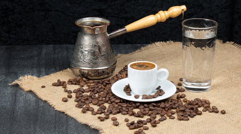 Hot coffee in Turkish Cooper and linen table. Greek Coffee Pot, Glass of water and Coffee beans dropped on wooden table.