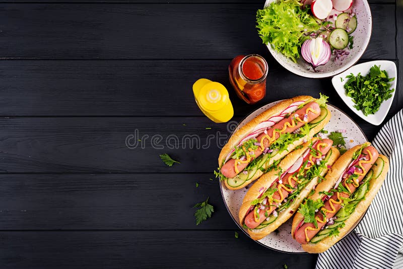 Hot dog with sausage, cucumber, radish and lettuce on dark wooden background.